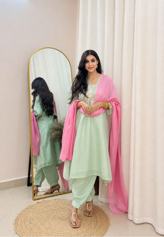 A woman models a light green traditional outfit with a pink dupatta, standing in front of a full-length mirror and white curtains in a well-lit room.