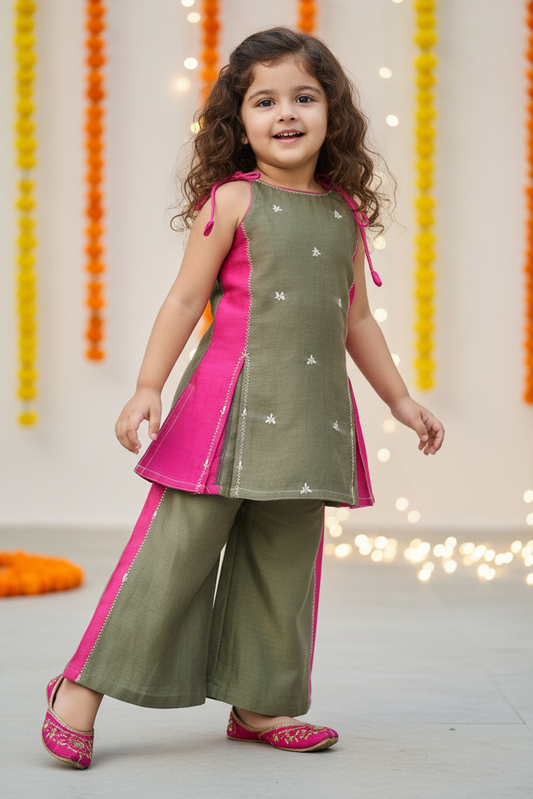 A young girl modeling a traditional Indian outfit consisting of an olive green kurta with pink side panels and silver star embroidery, paired with matching wide-leg trousers and pink shoes, set against a decorated background with marigold garlands.
