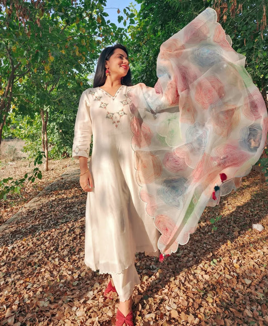 Woman in a white dress with a floral dupatta standing outdoors among trees.