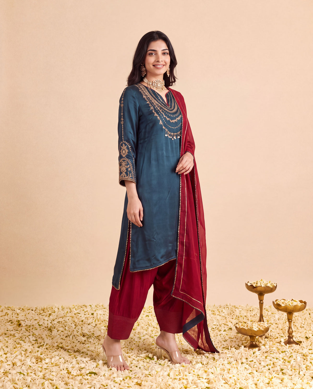A woman models a traditional Indian salwar kameez suit set consisting of a slate blue kurta with gold embroidery, maroon pants, and a maroon dupatta, standing on a bed of flower petals against a beige background.