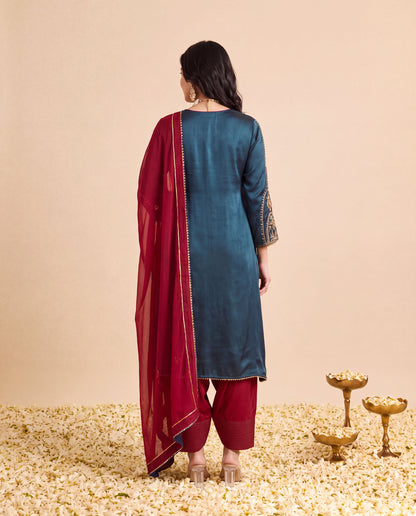 A woman stands with her back to the camera wearing a dark teal kurta with gold embroidery on the sleeves, matching maroon pants, and a deep maroon dupatta draped over her shoulder. She is standing on a bed of white flower petals against a solid beige background, with three small gold decorative lamps on the right.