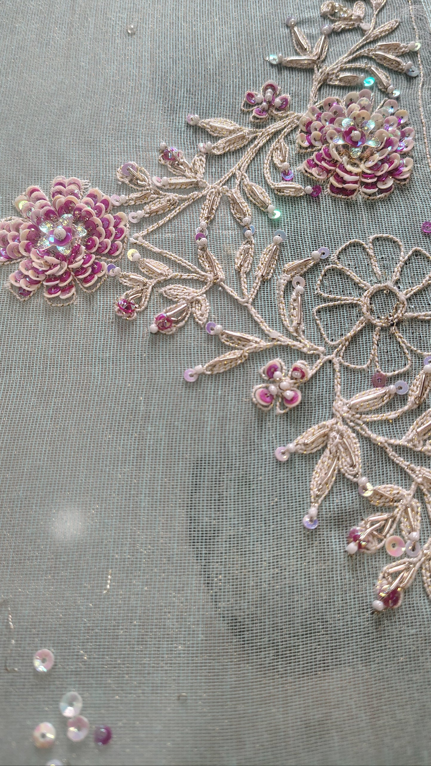 Close-up of a sheer gray fabric decorated with pink floral embroidery and metallic gold leafy vines embellished with small iridescent beads.