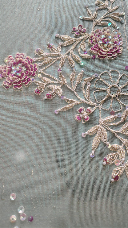 Close-up of a sheer gray fabric decorated with pink floral embroidery and metallic gold leafy vines embellished with small iridescent beads.