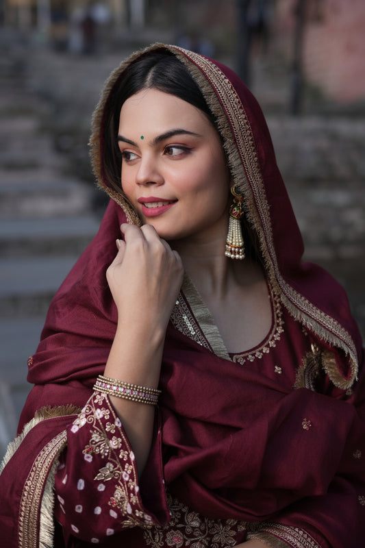 A woman wearing a dark green traditional outfit with an embroidered hooded shawl, gold-toned jewelry, and a bindi, posed against a softly blurred outdoor stone staircase background.