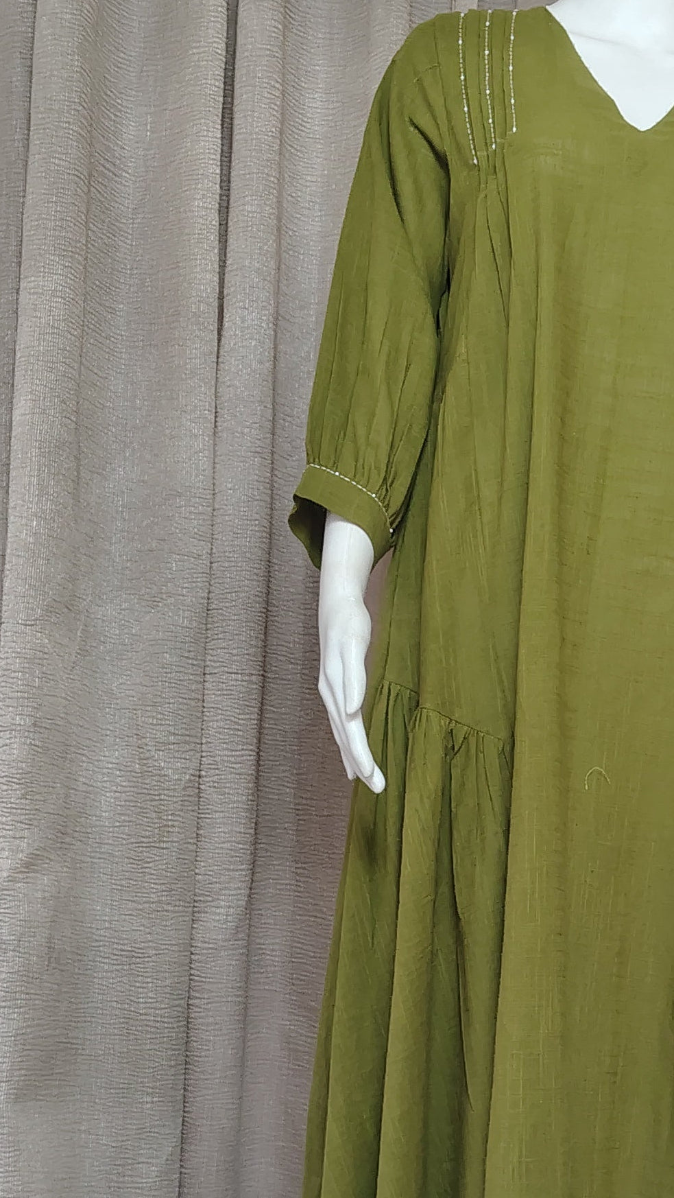 An olive green long-sleeve dress displayed on a white mannequin, set against a backdrop of draped gray fabric.