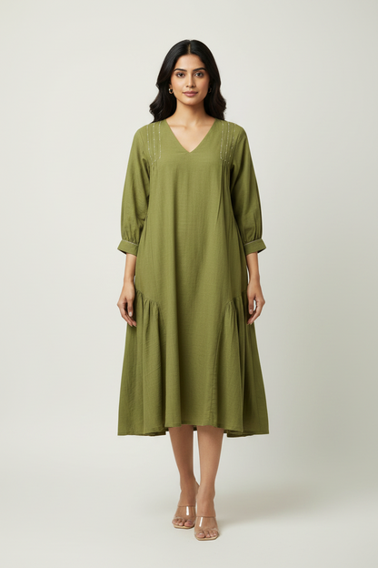 A woman modeling a loose olive green midi dress with three-quarter sleeves, a V-neckline, and subtle embroidered details, set against a plain light gray studio background.
