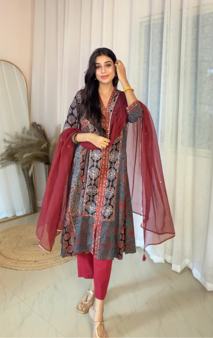 A woman models a traditional Indian salwar kameez suit set in a bright room with sheer white curtains. The outfit features a gray and red patterned kameez jacket with a red border, matching red trousers, and a deep red dupatta.