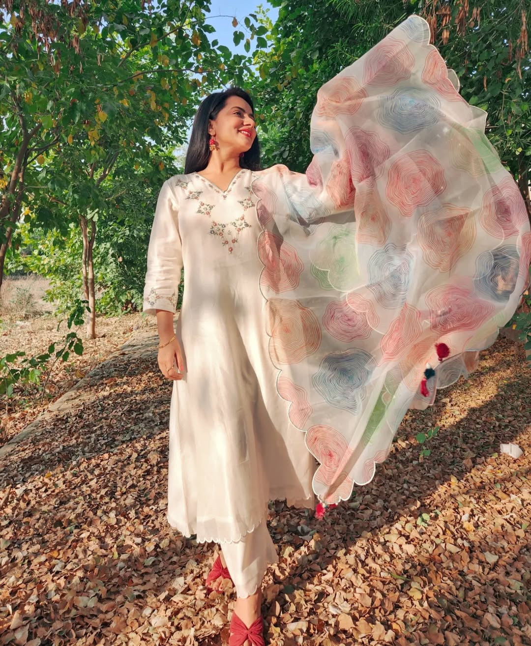 Woman in a white dress with a floral dupatta standing outdoors among trees.